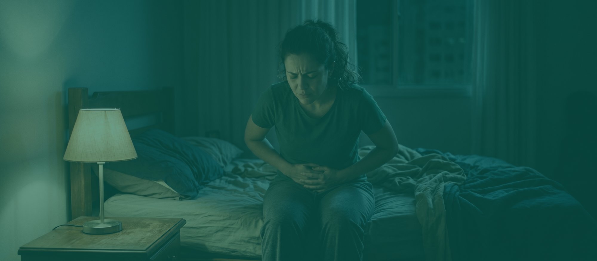 Mulher curvada na beira da cama à noite, mãos no baixo ventre e expressão de dor, quarto iluminado só por um abajur