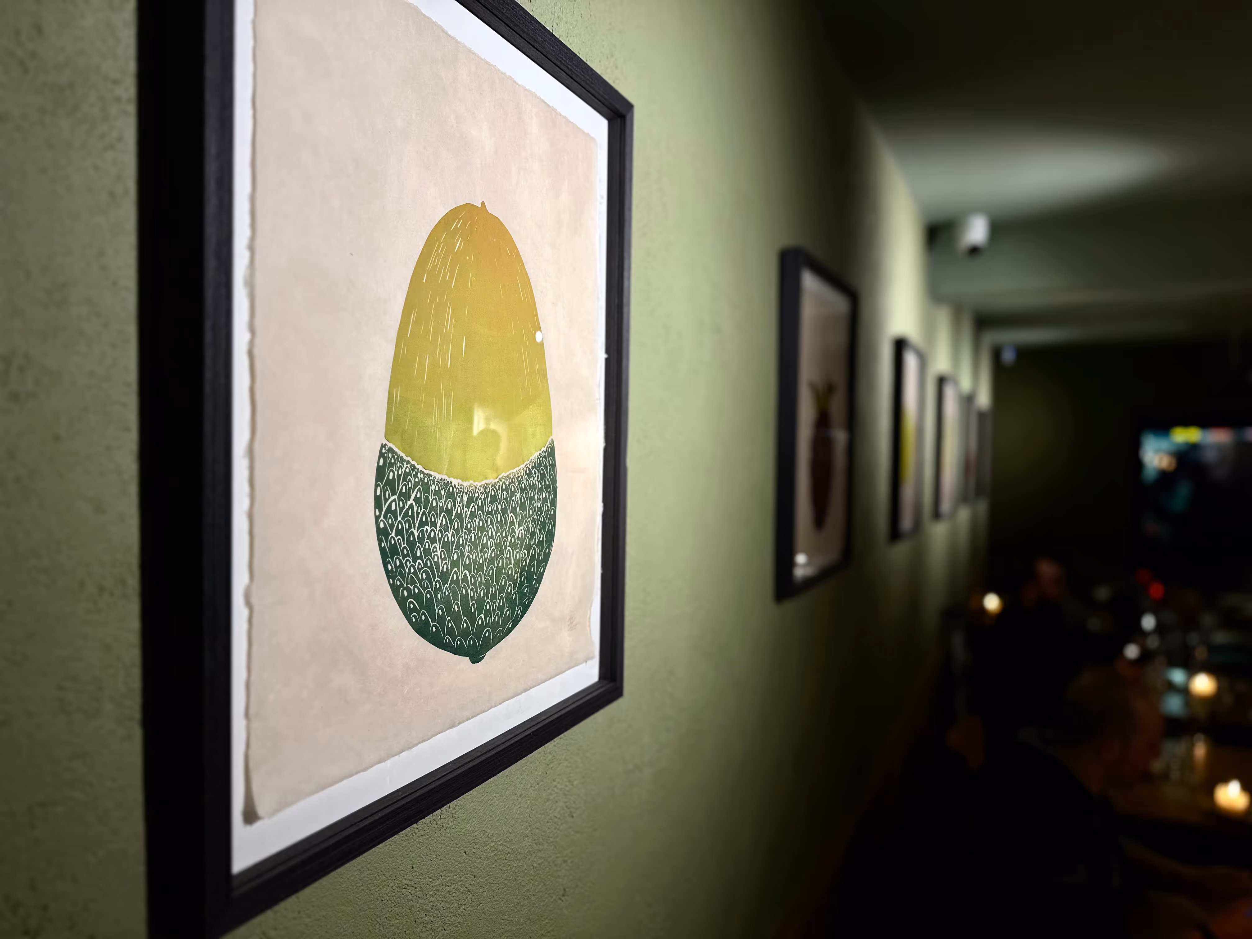 Framed abstract art with a yellow top and patterned green bottom on a green wall in a dim room.