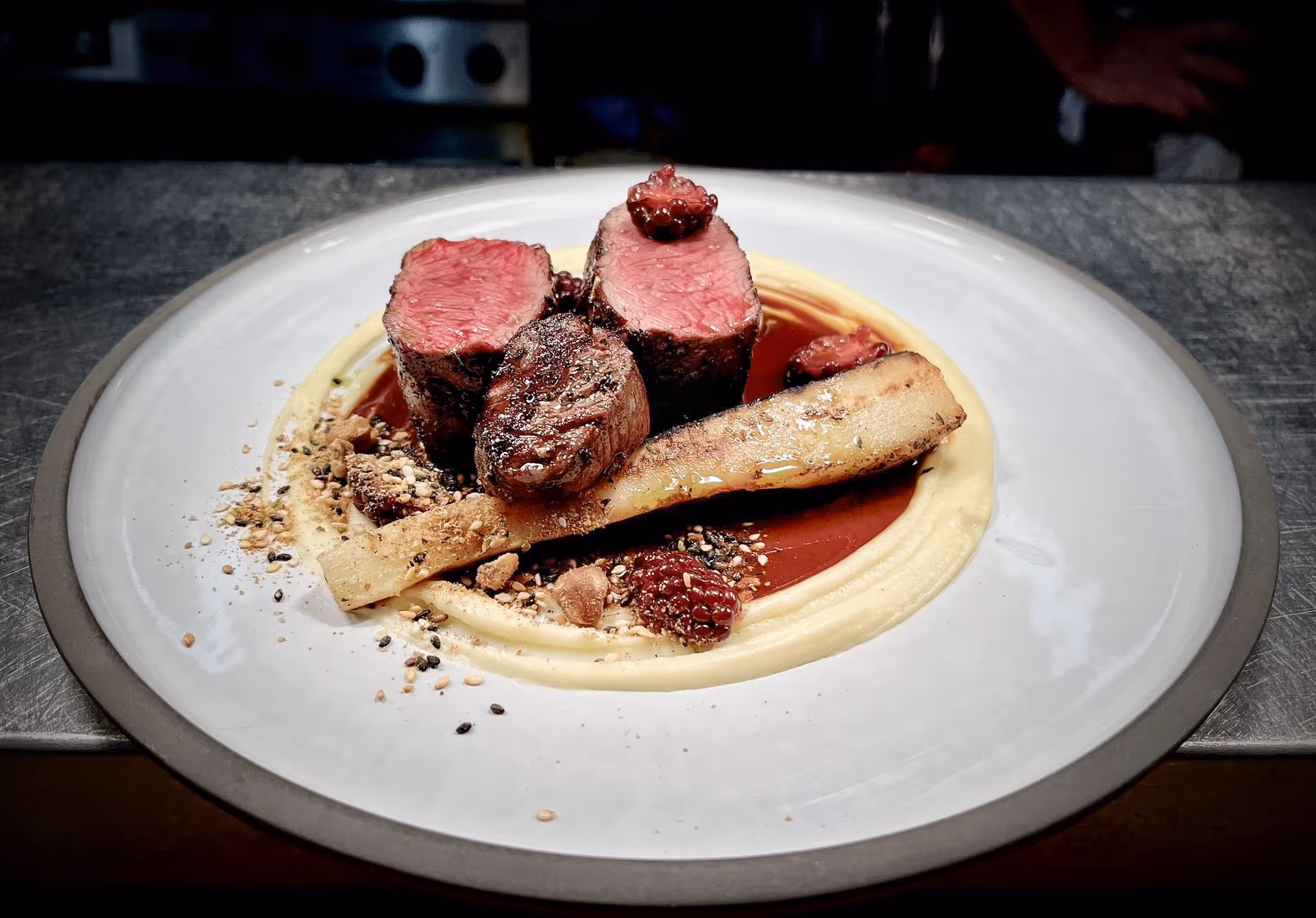 Three slices of medium-rare steak on creamy puree with sauce, raspberries, and seeds on a white plate.