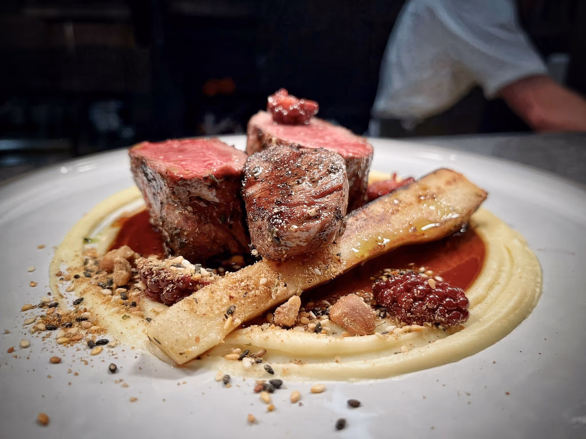 Plate with medium-rare steak pieces on creamy puree, sauce, nuts, and berries garnished with a crispy element.