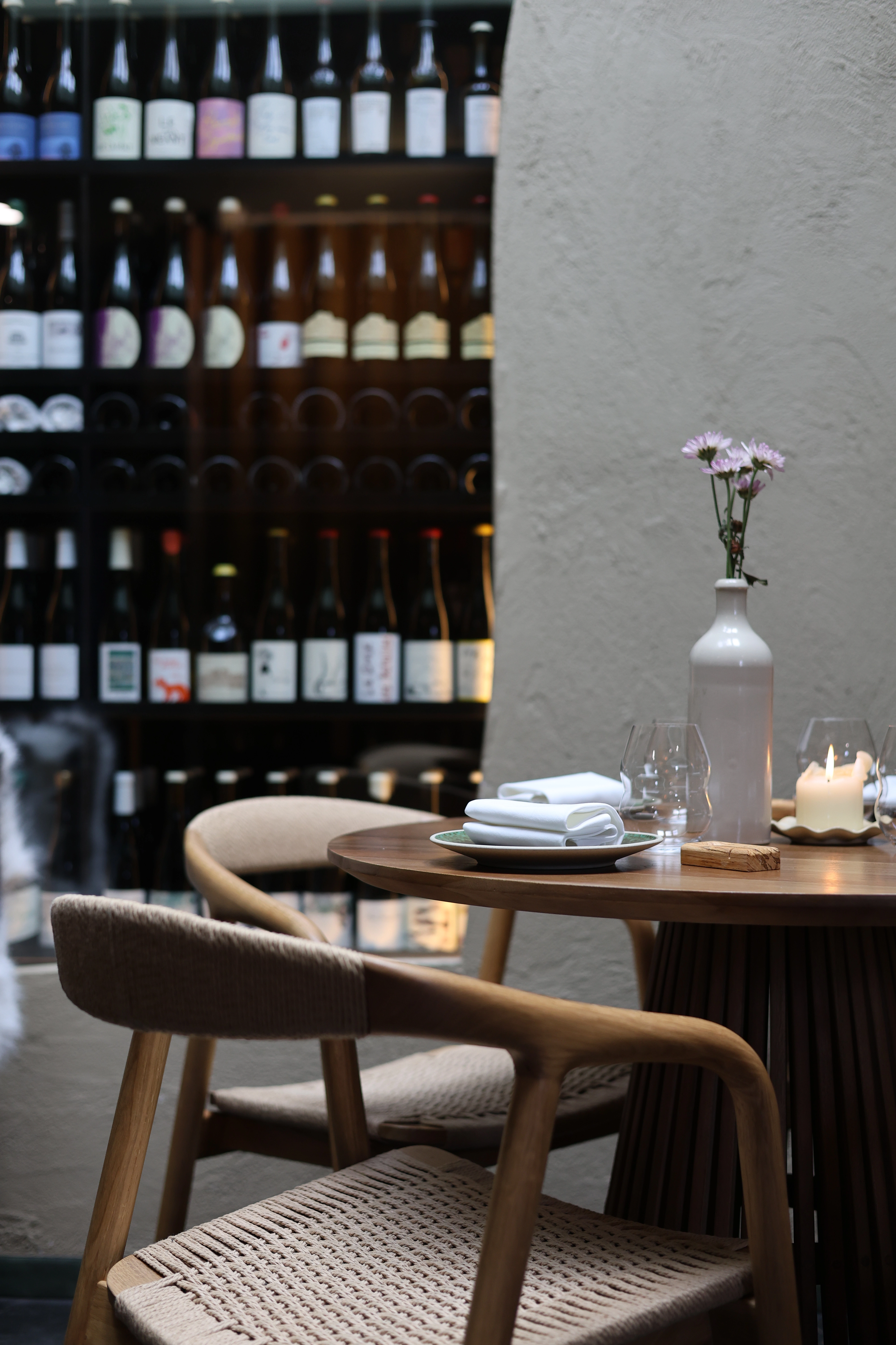 Round wooden table with plates, glasses, a vase with flowers, candles, and woven chairs near wine bottles.
