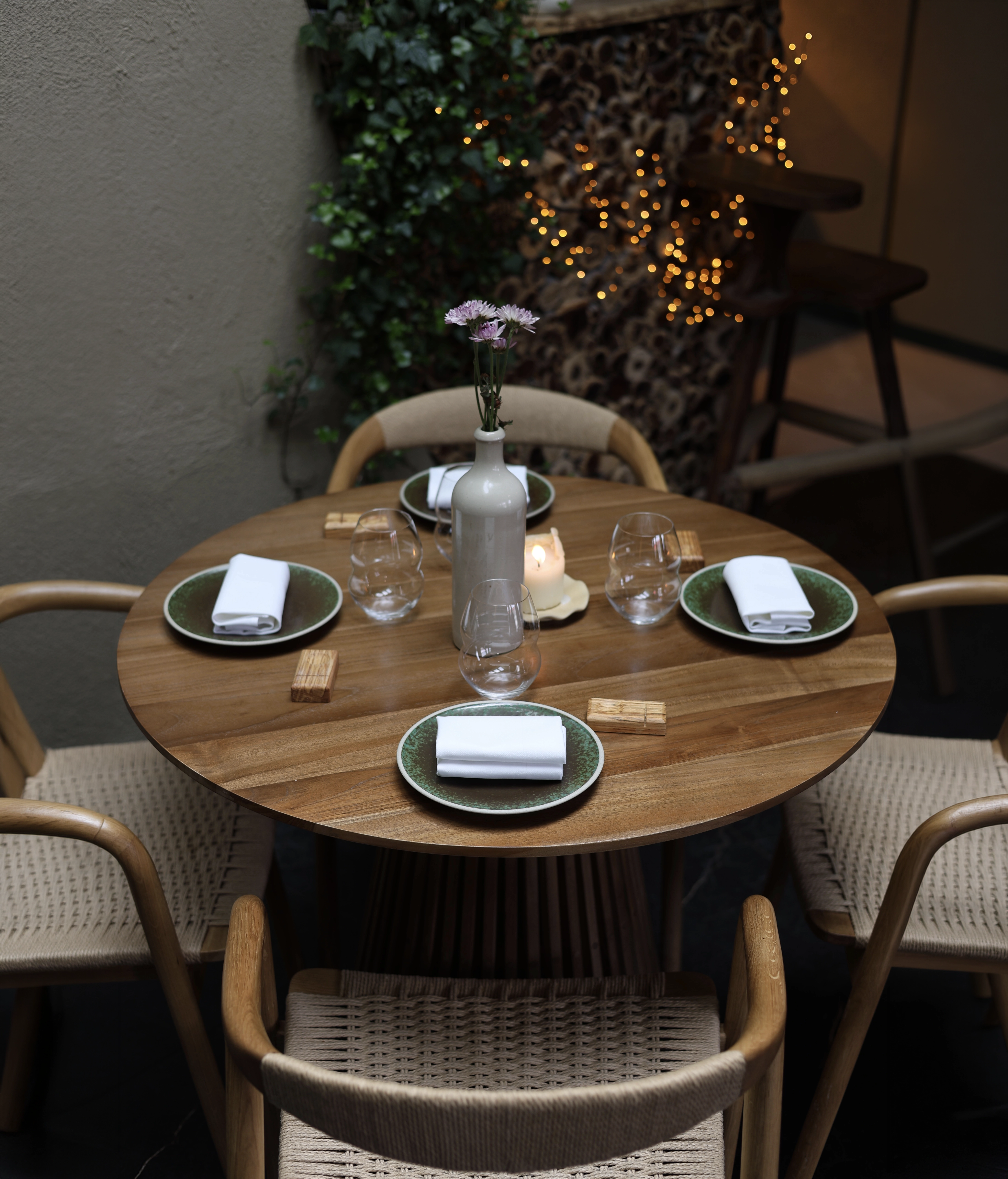 Round wooden table set for four with green plates, folded white napkins, glasses, and vase with flowers.