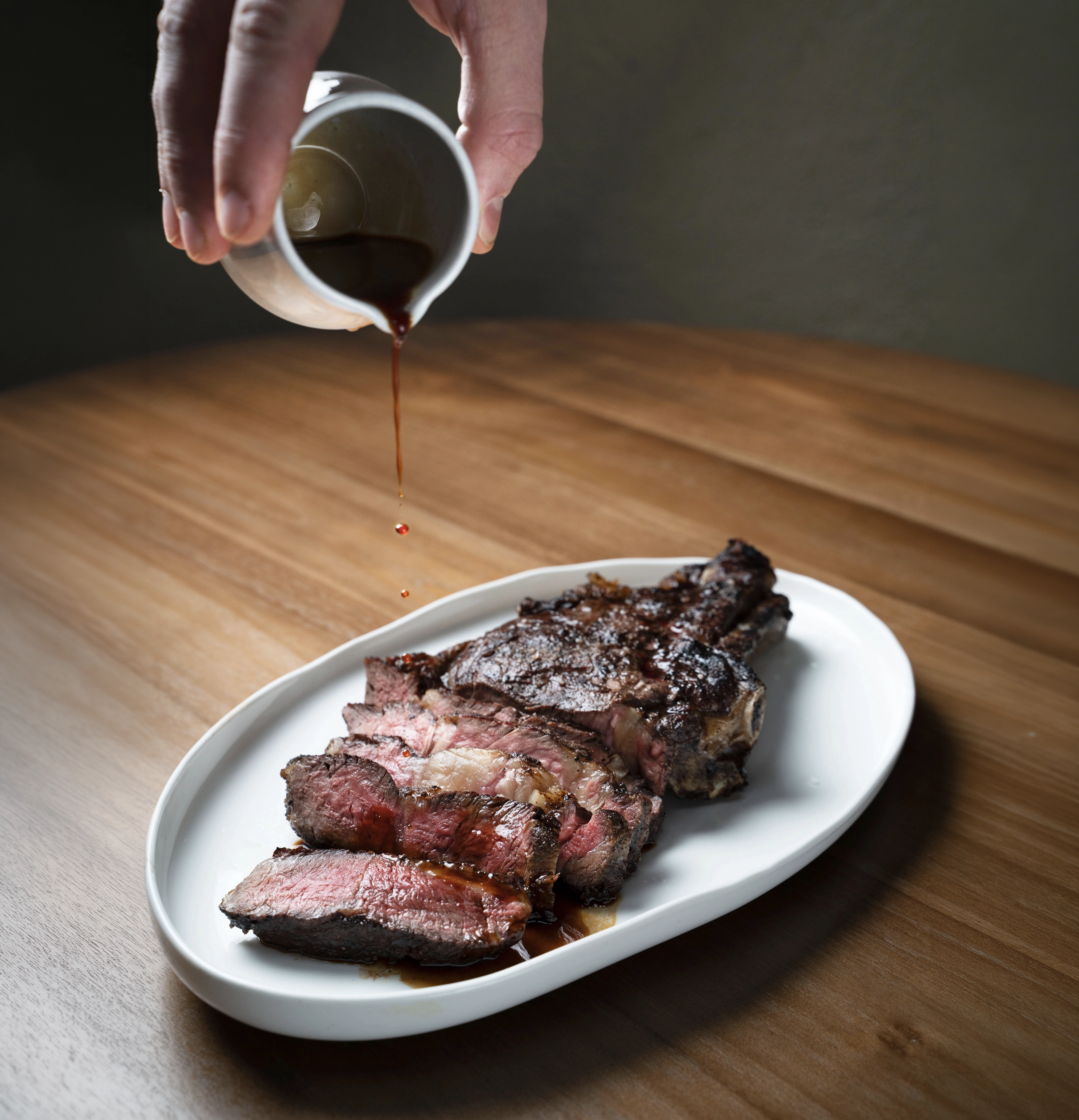 Sauce being poured over sliced medium-rare steak on a white plate on a wooden surface.