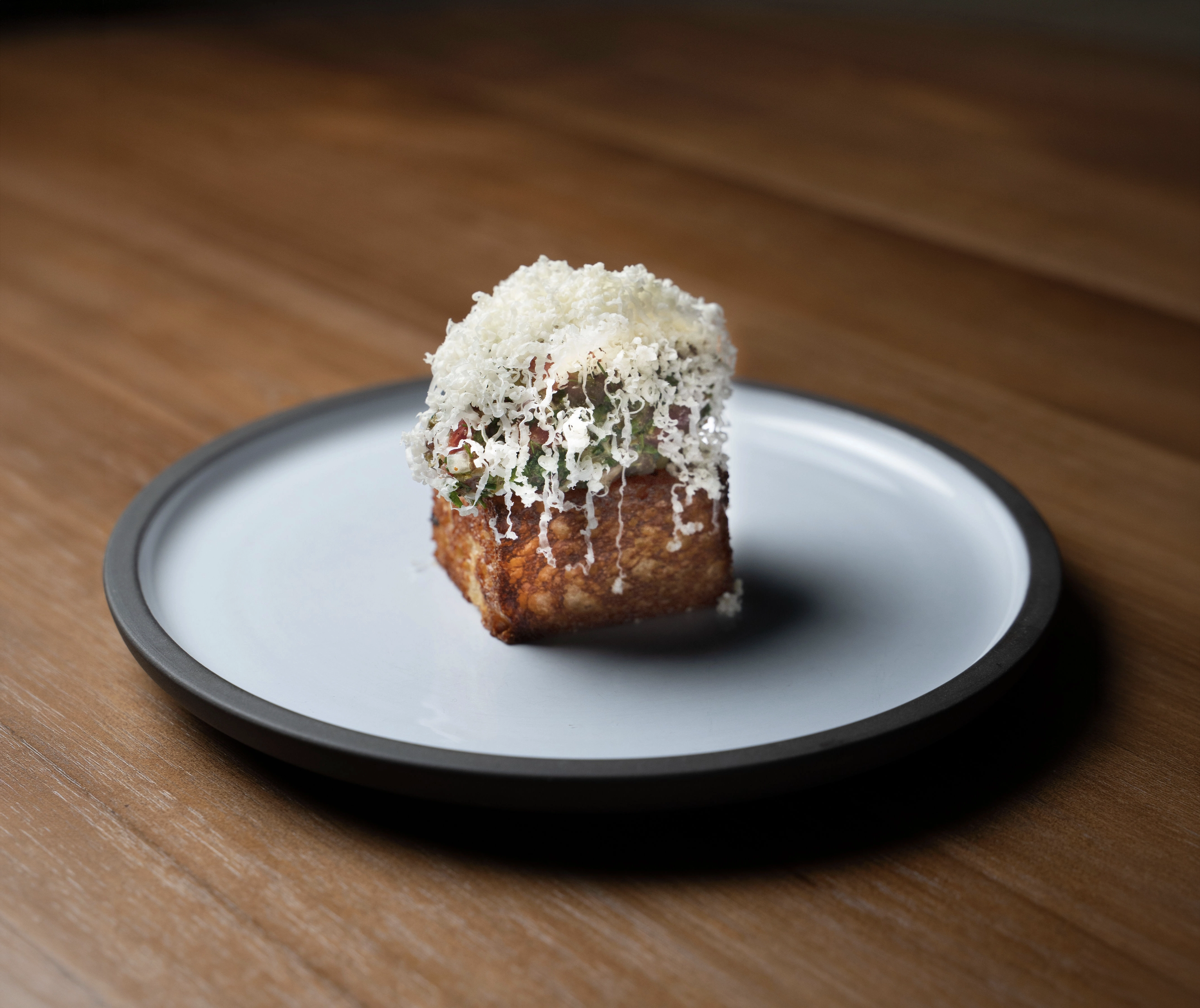 Square piece of toasted bread topped with herbs and shredded white cheese on a gray-rimmed plate.