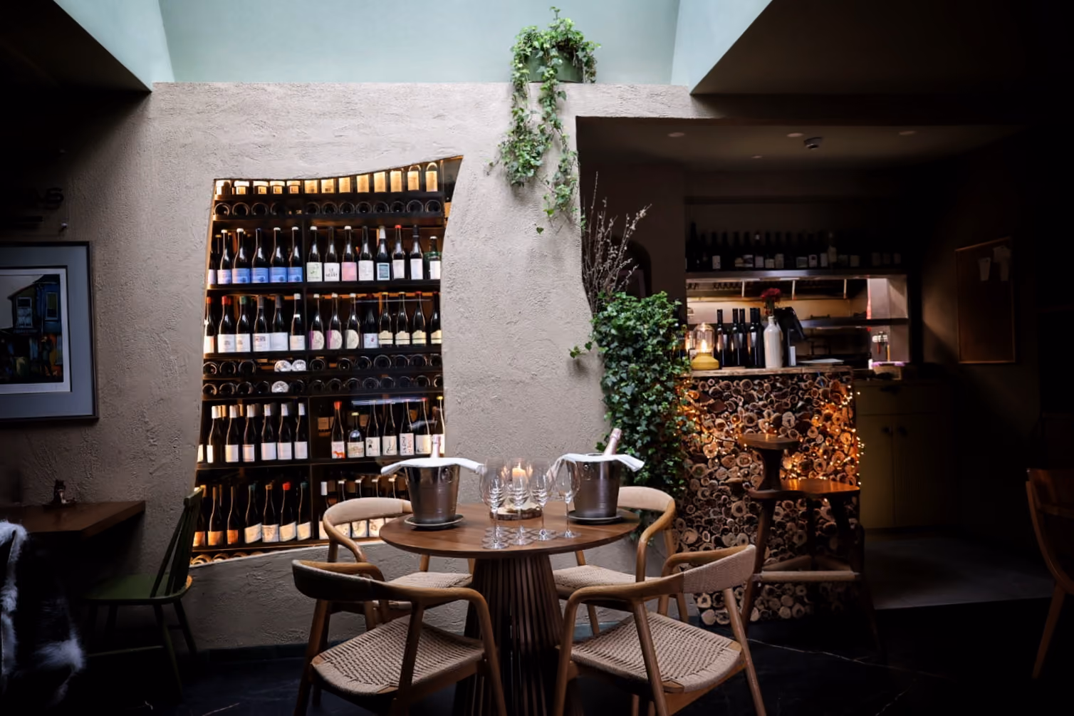 Cozy restaurant corner with a round table, four chairs, wine bottles on shelves, and green plants.