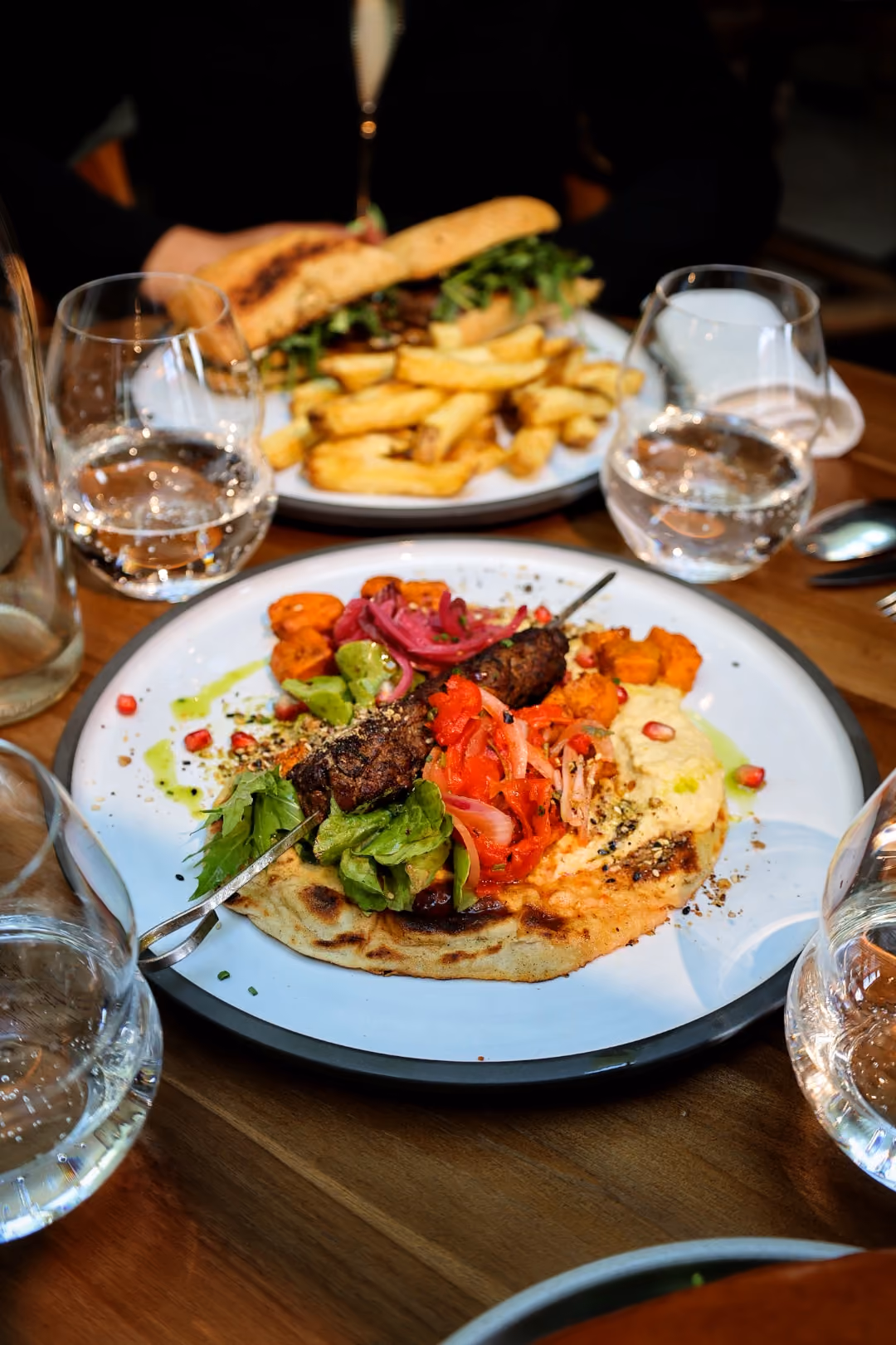 Plate with grilled meat skewer, roasted vegetables, greens, and hummus on flatbread on a wooden table.