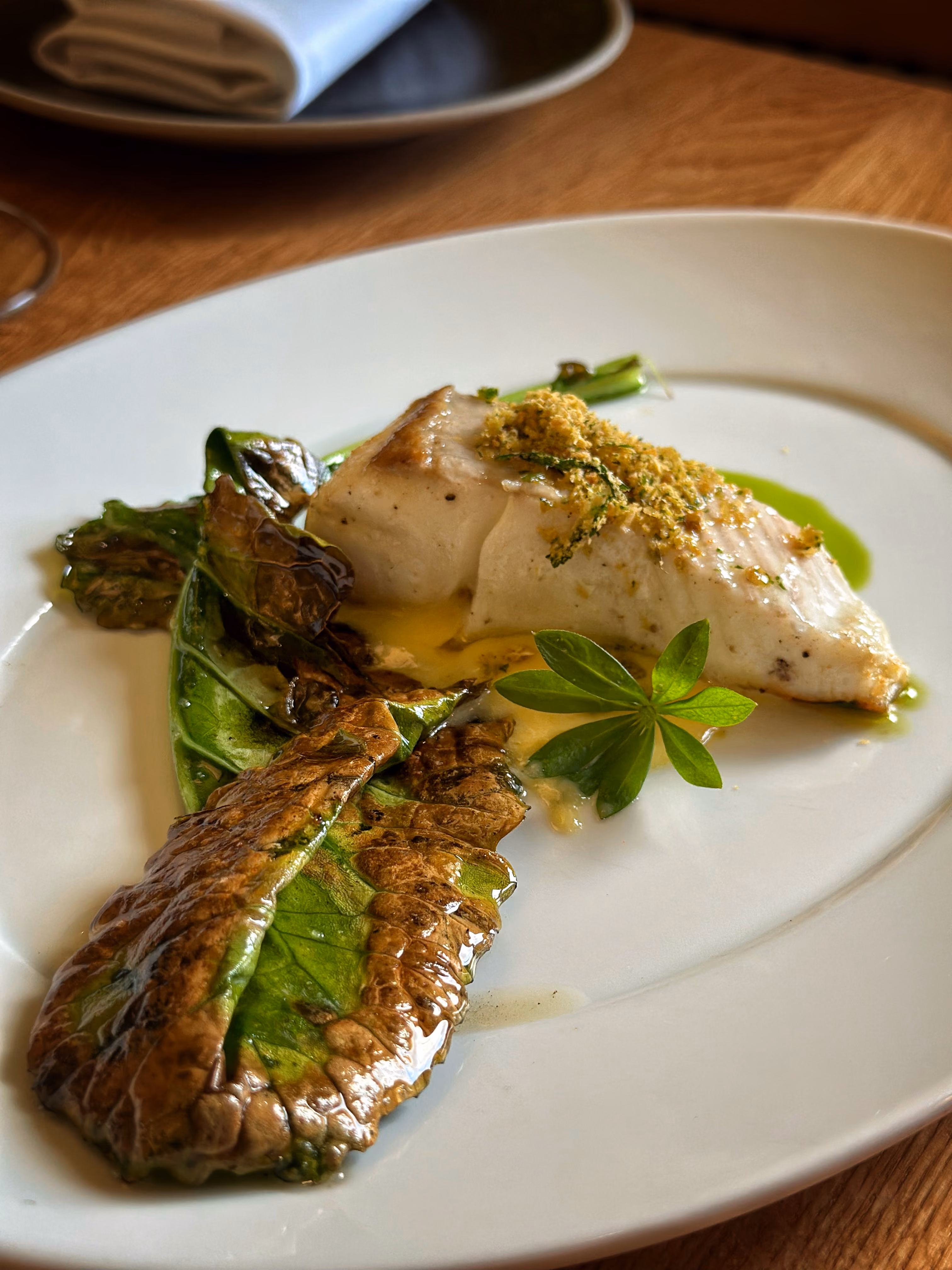 Gourmet plate with seared fish topped with herb crumb, wilted greens, and a small fresh leaf garnish.