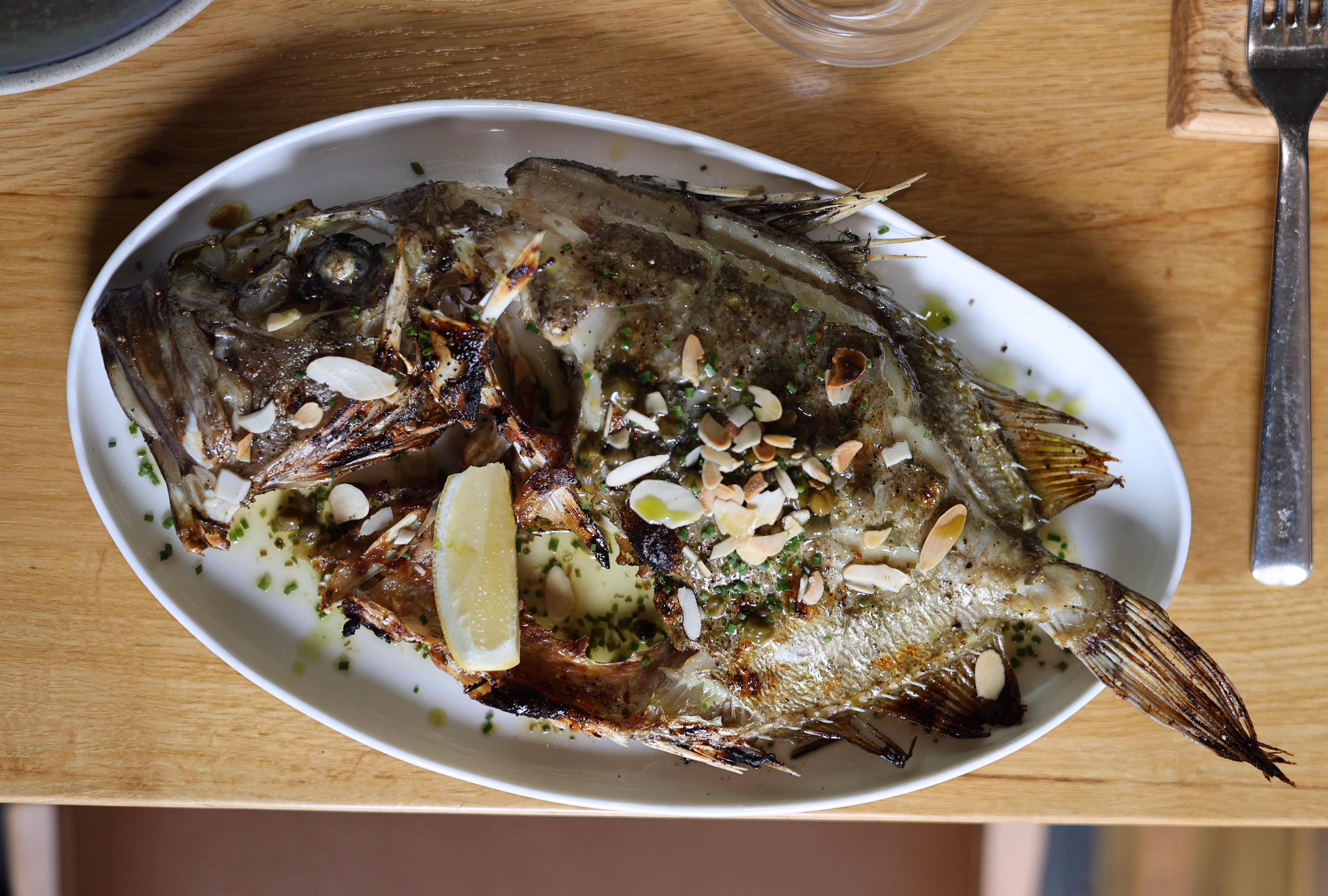 Grilled whole fish served on a white plate with lemon wedge and sprinkled with herbs and sliced almonds.