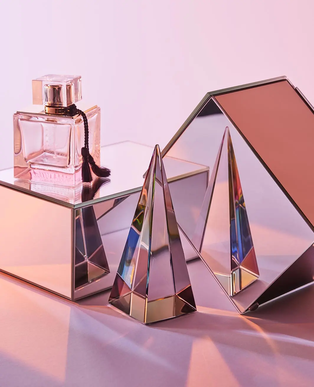 Pink perfume bottle on reflective mirrored cube beside a glass pyramid and reflective triangular prism.