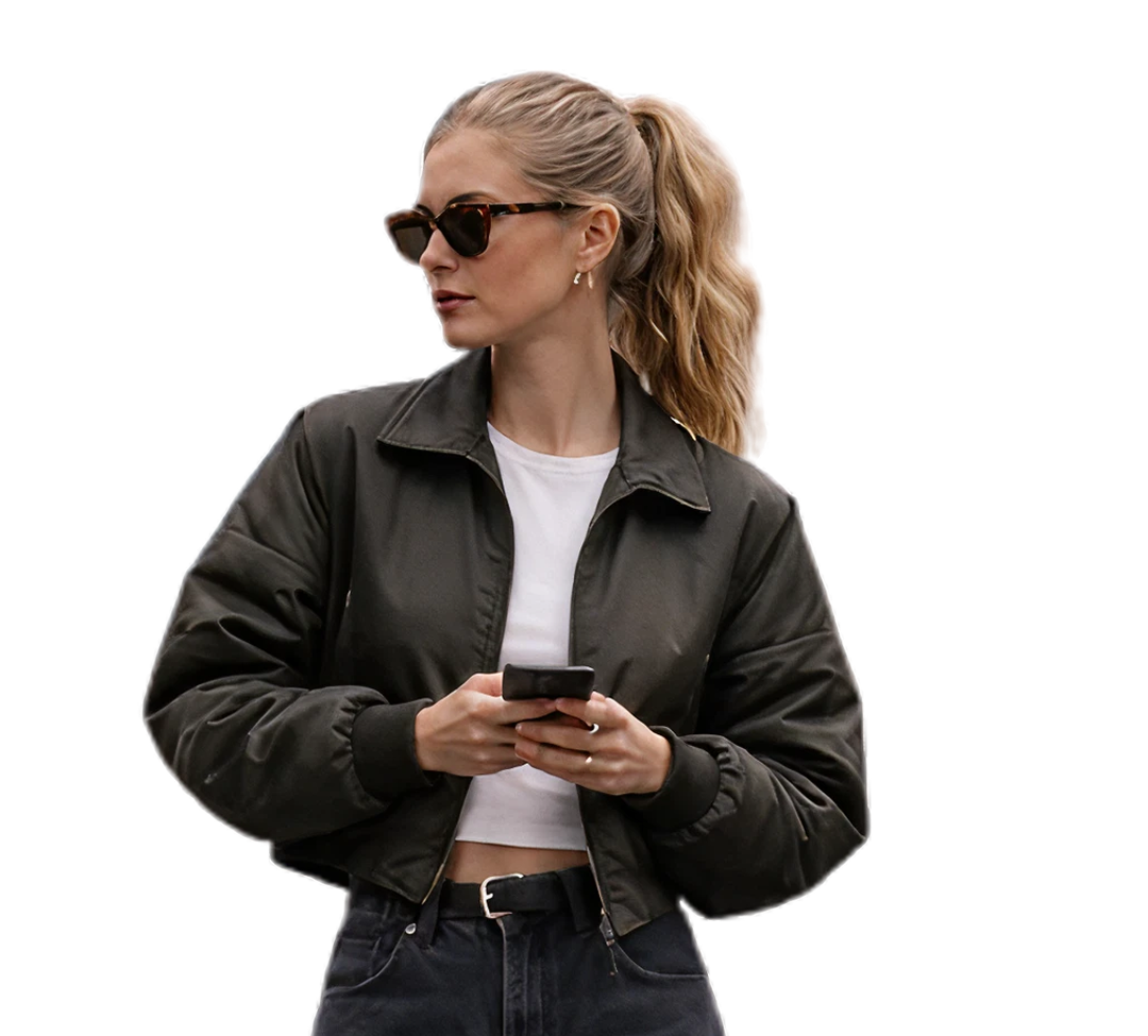 Woman with blonde hair in a ponytail wearing sunglasses, a black jacket, white crop top, and dark pants holding a smartphone.