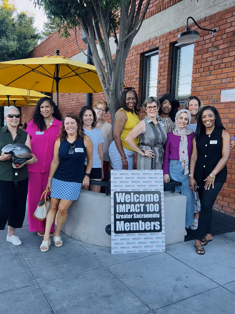 Group of diverse women smiling outdoors around a sign welcoming IMPACT 100 Greater Sacramento members.