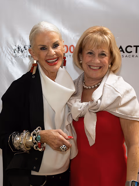 Two smiling women posing together, one in black with white scarf, the other in red with pearl necklace.