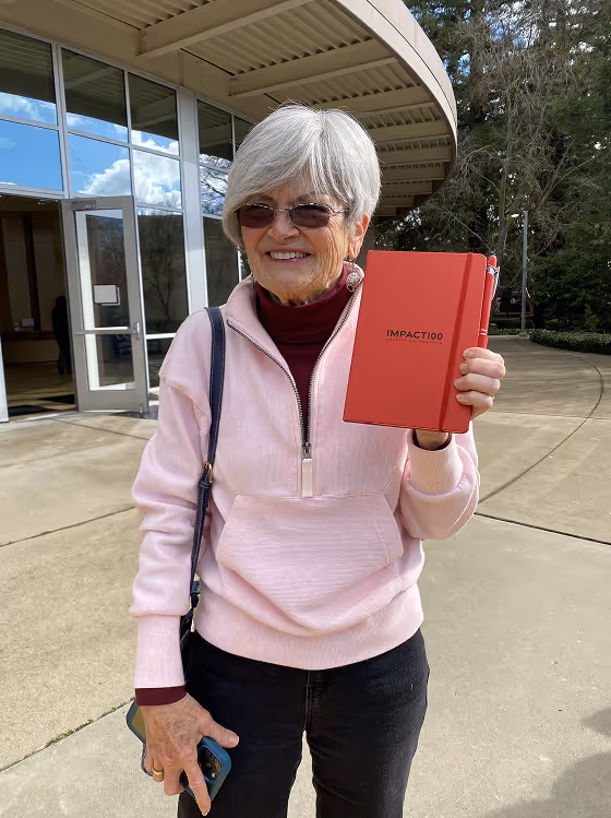 Smiling older woman with silver hair holding a red IMPACT100 notebook outdoors.