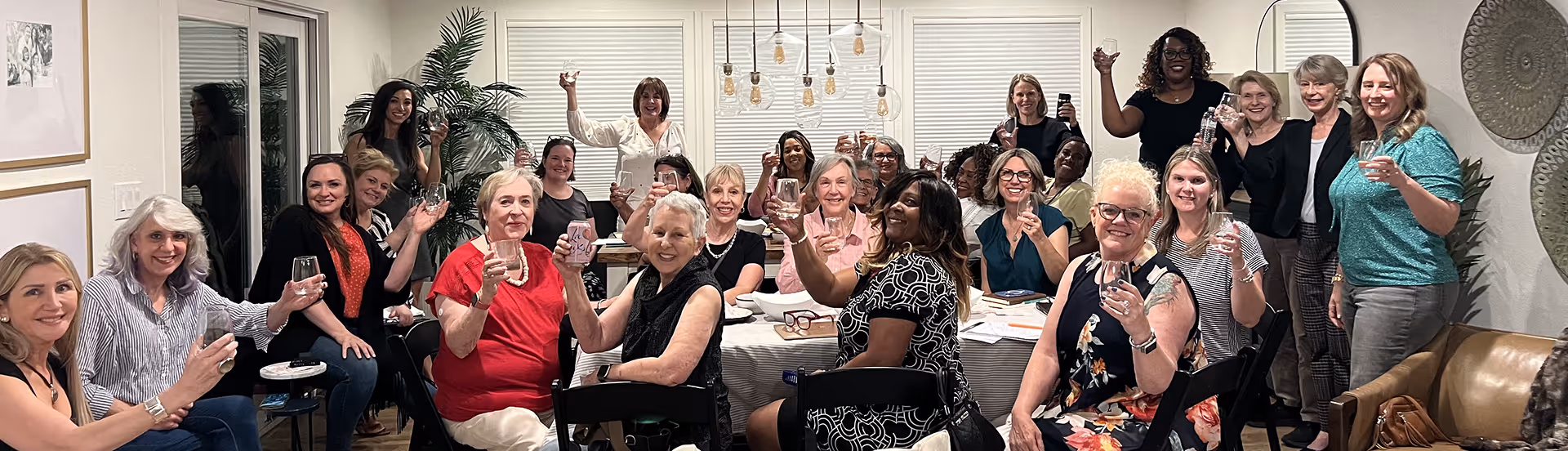 Group of women raising glasses in a toast around a dining table in a well-lit living room.