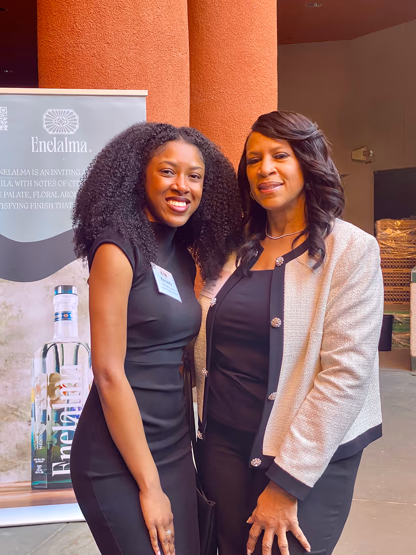 Two women standing and smiling, one in a black dress and the other in a white jacket over black dress.