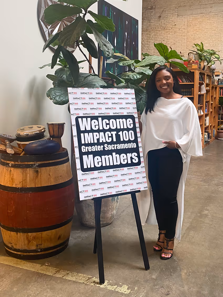 Woman in white top and black pants smiles beside a sign welcoming IMPACT 100 Greater Sacramento members.