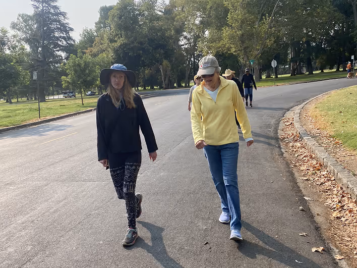 Two women walking on a paved road in a park with trees and grass around.