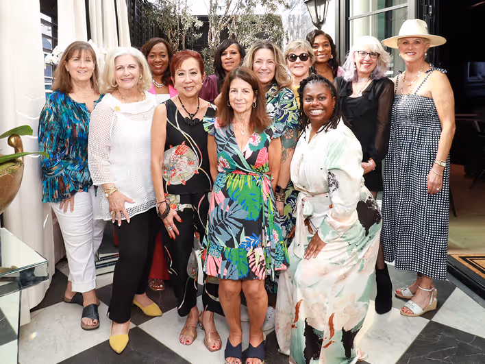 Group of 12 diverse women posing together indoors, smiling and dressed in colorful, stylish outfits.