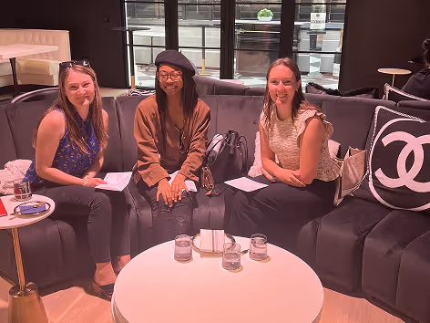 Three smiling women sitting on a black couch around a white round table with glasses and napkins.