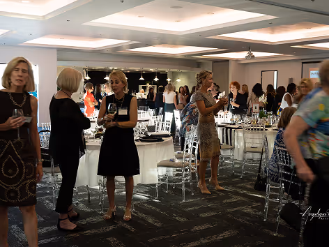 Women socializing and drinking wine at a formal indoor event with round tables and clear chairs.