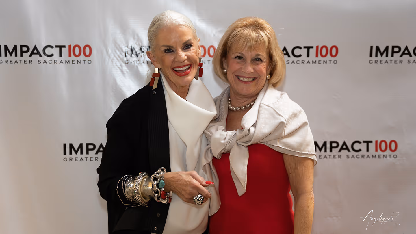 Two smiling older women pose in front of an Impact100 Greater Sacramento backdrop at an event.