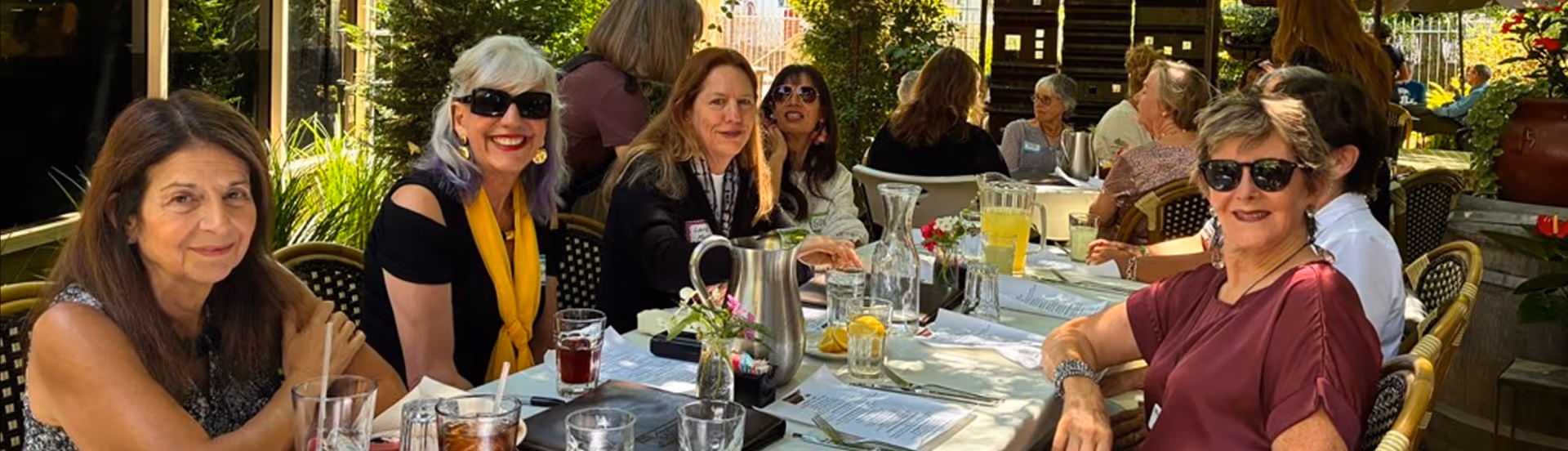 Group of women dining outdoors at a long table with drinks and menus in daylight.