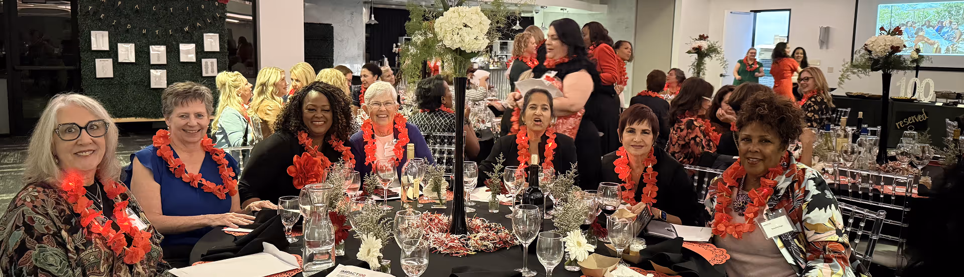 Group of women wearing red leis seated around a table with flowers and wine glasses at an event.