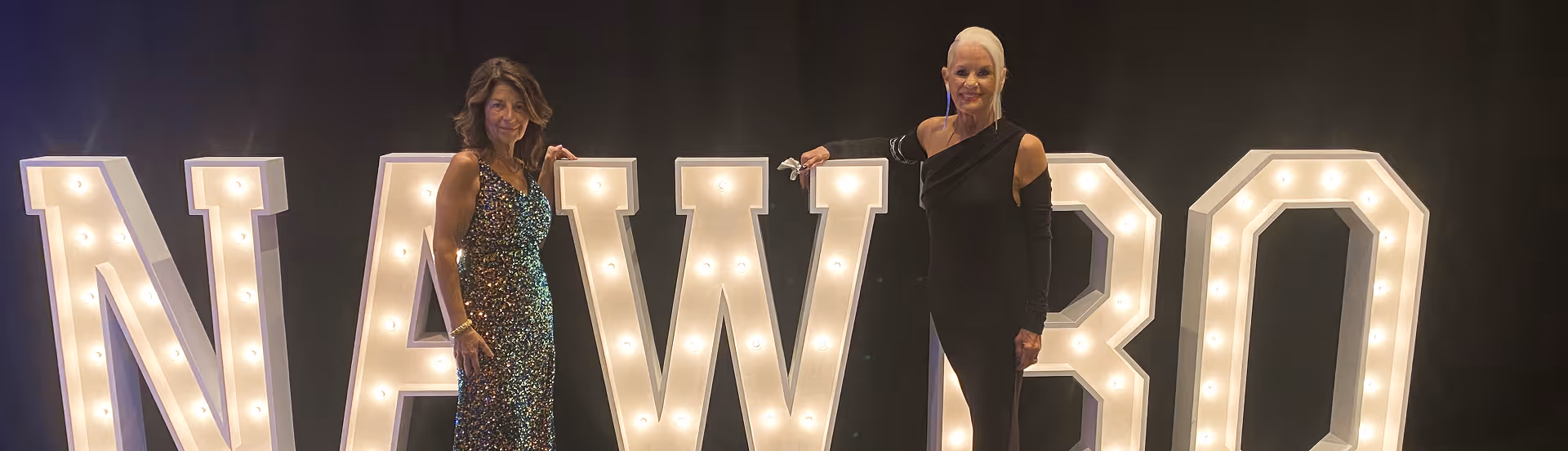 Two women in formal dresses standing beside large illuminated letters spelling NAWBO.