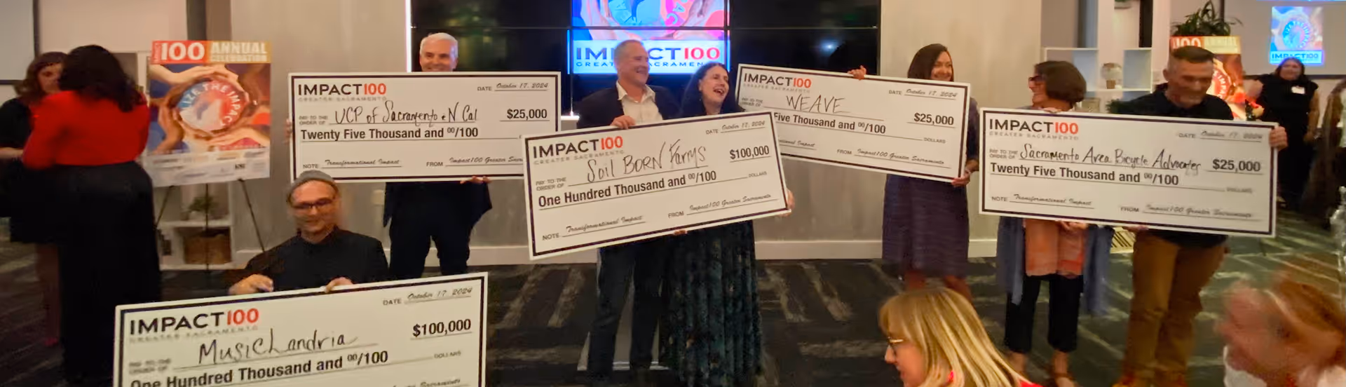 Group holding large Impact100 grant checks ranging from $25,000 to $100,000 in an event room.