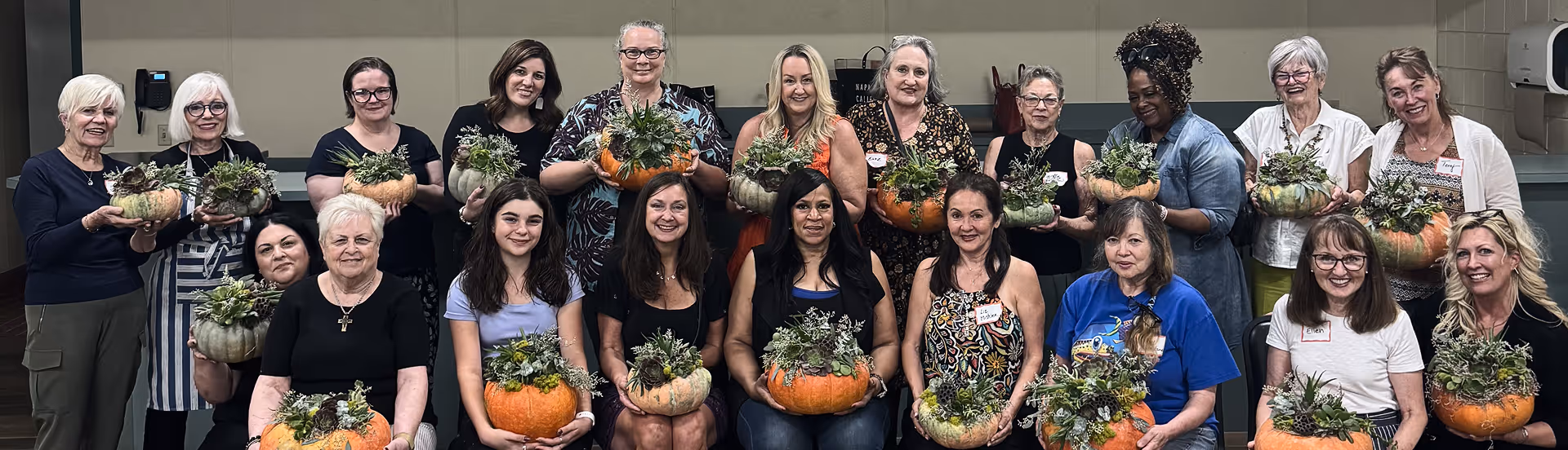 Group of women holding pumpkin succulent planters, smiling indoors against beige wall.