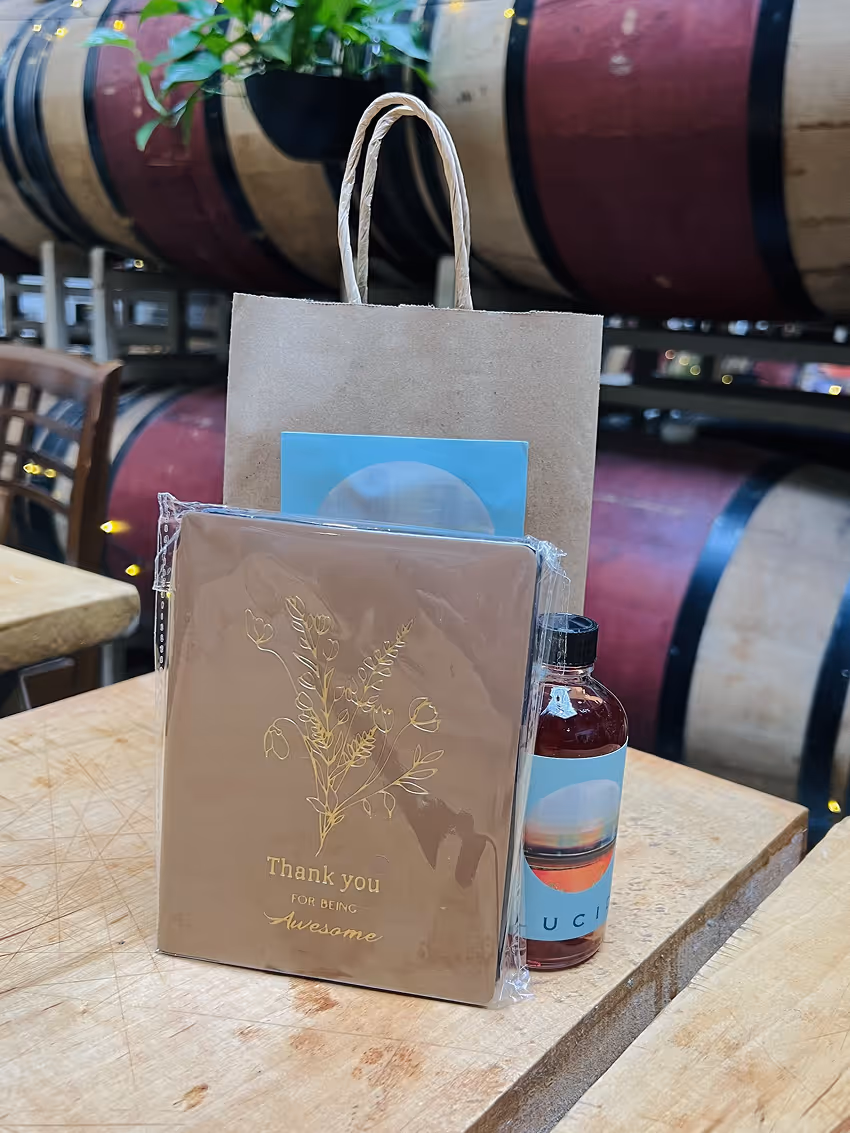 Gift bag with a thank you notebook and a small bottle on a wooden table with wine barrels in background.