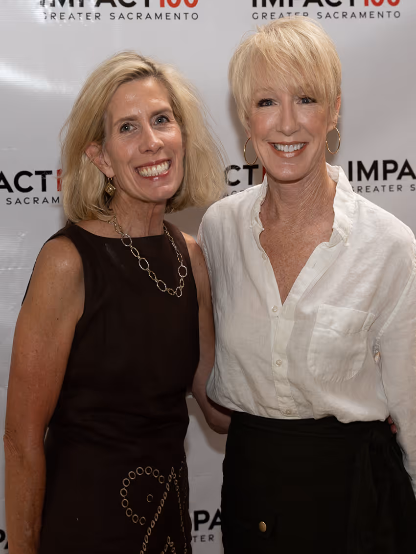 Two smiling women standing side by side in front of an Impact100 Greater Sacramento backdrop.