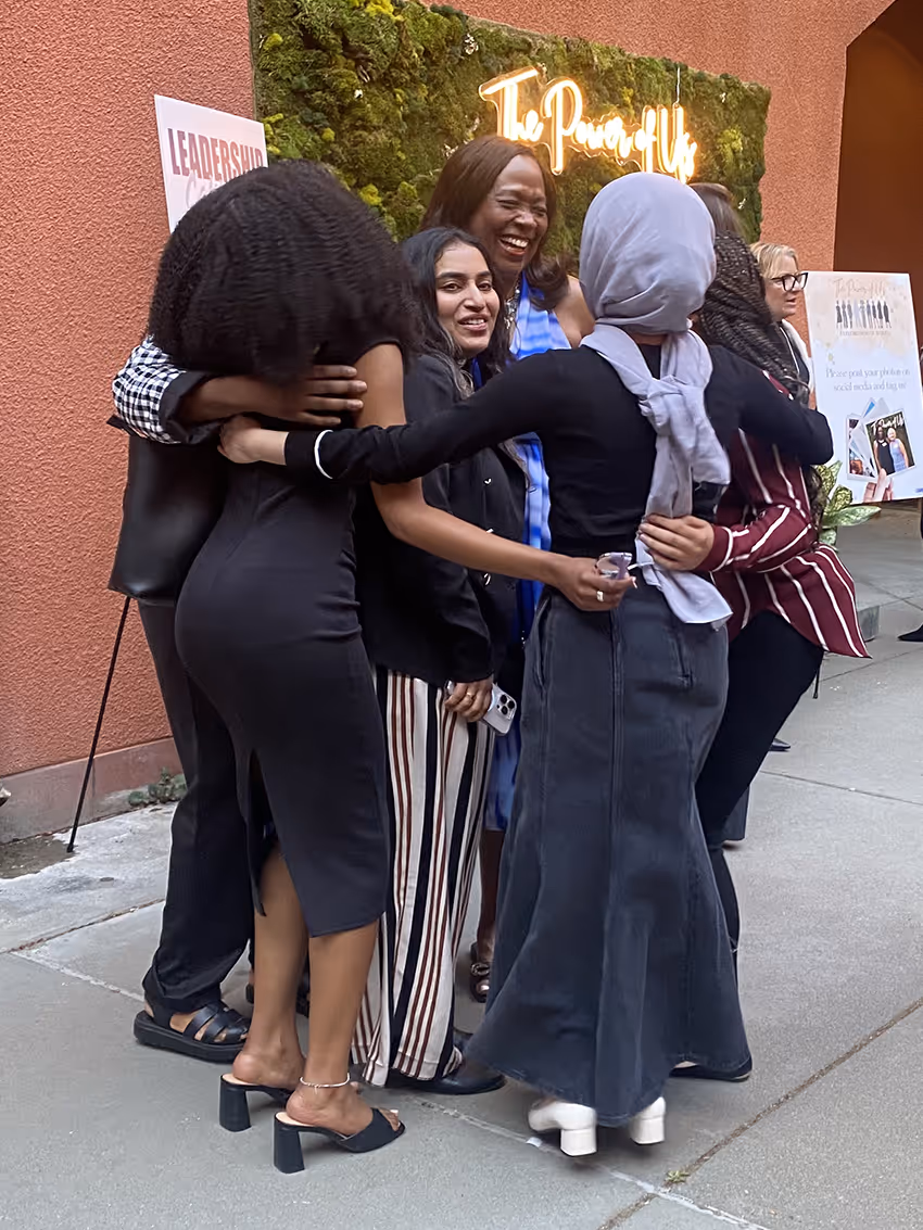 A group of diverse women smiling and hugging outside near a wall with a neon sign reading 'The Power of Us'.