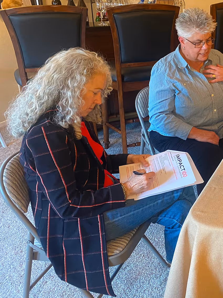 Woman with curly hair sitting, writing on an Impact100 grant eligibility form at a table.