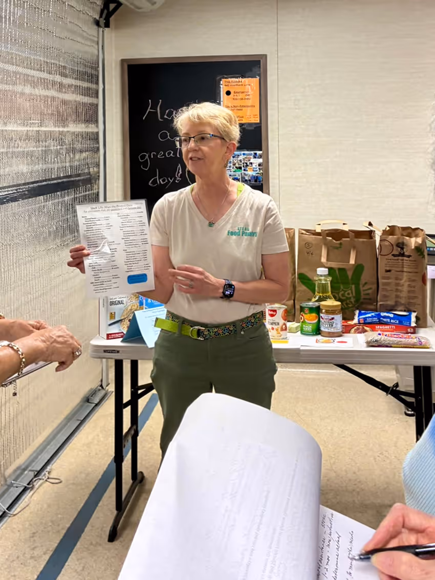 Volunteer in AFCB Food Pantry shirt holds a paper, talking to people near a table with groceries.