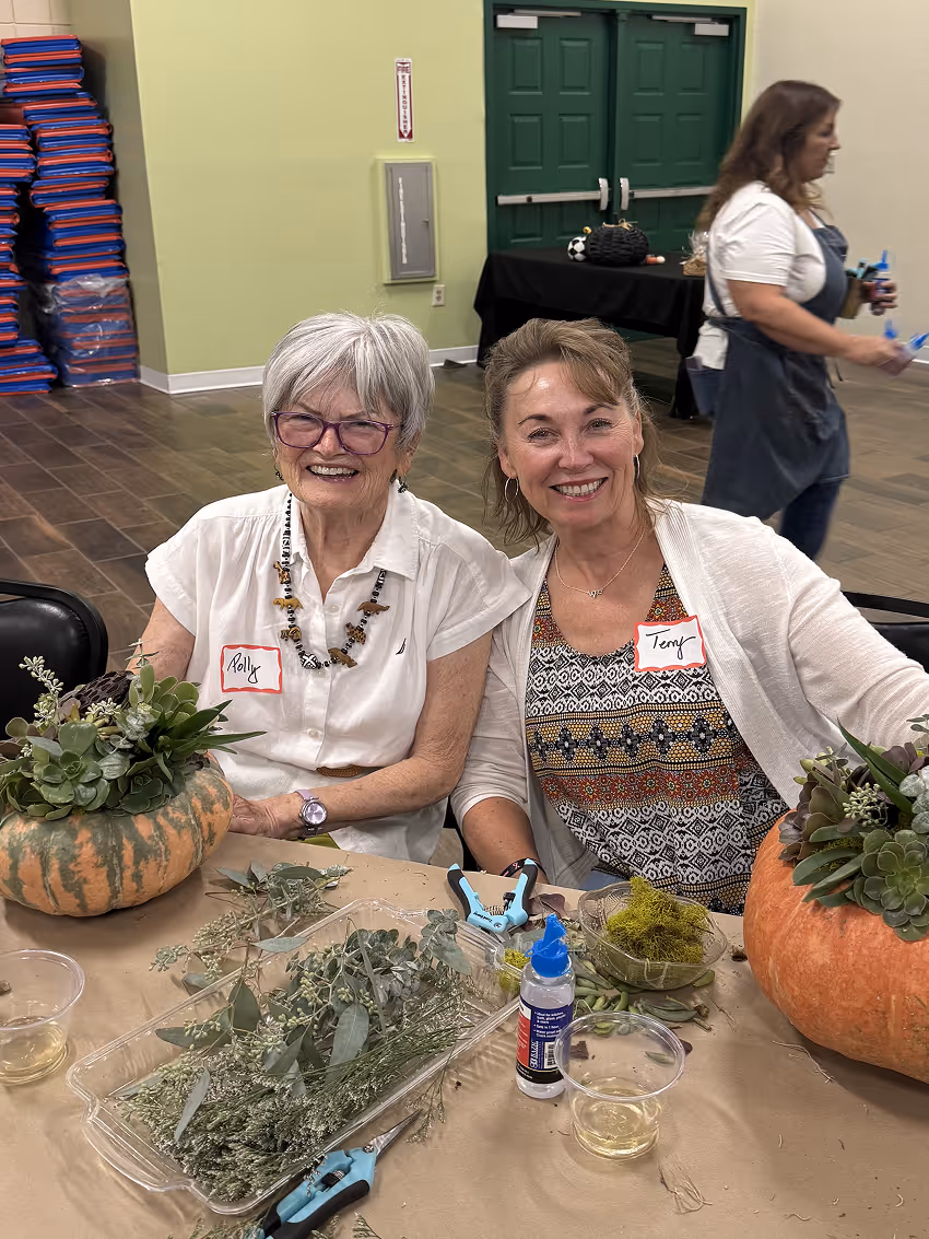 Two smiling women with name tags sit at a table arranging plants in pumpkins indoors.
