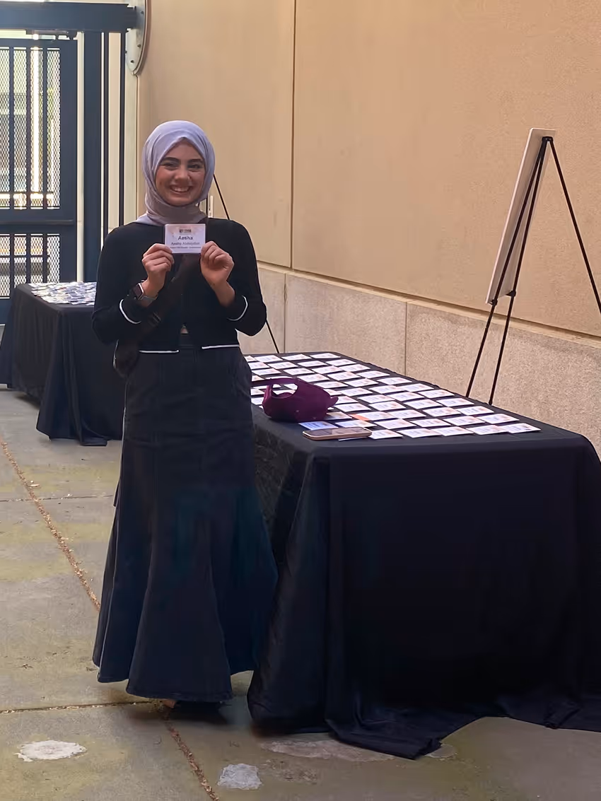 Woman with a lavender headscarf holding a name badge standing by tables with many badges.