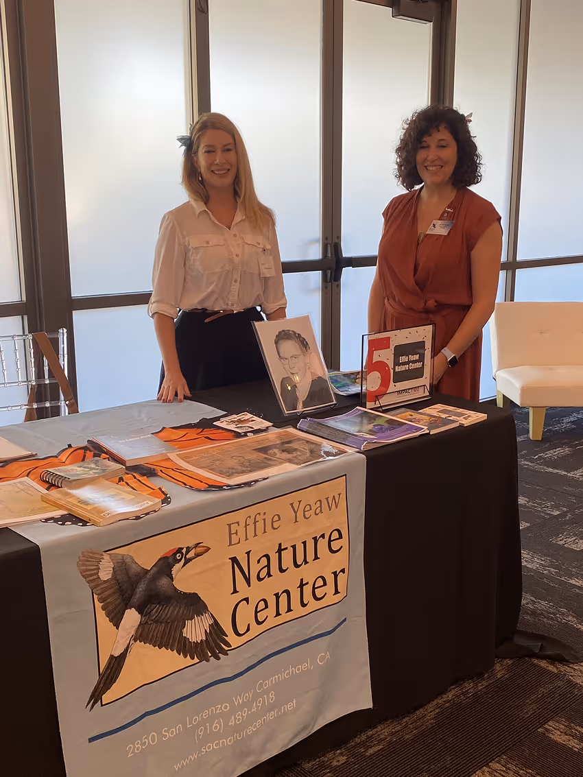 Two women standing behind a table promoting Effie Yeaw Nature Center with brochures and a portrait.