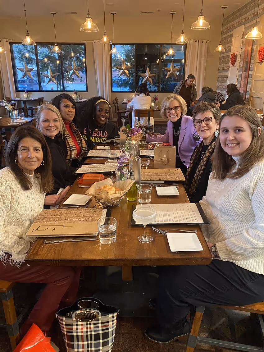 Seven women sitting around a wooden table with menus, drinks, and bread baskets in a decorated restaurant.