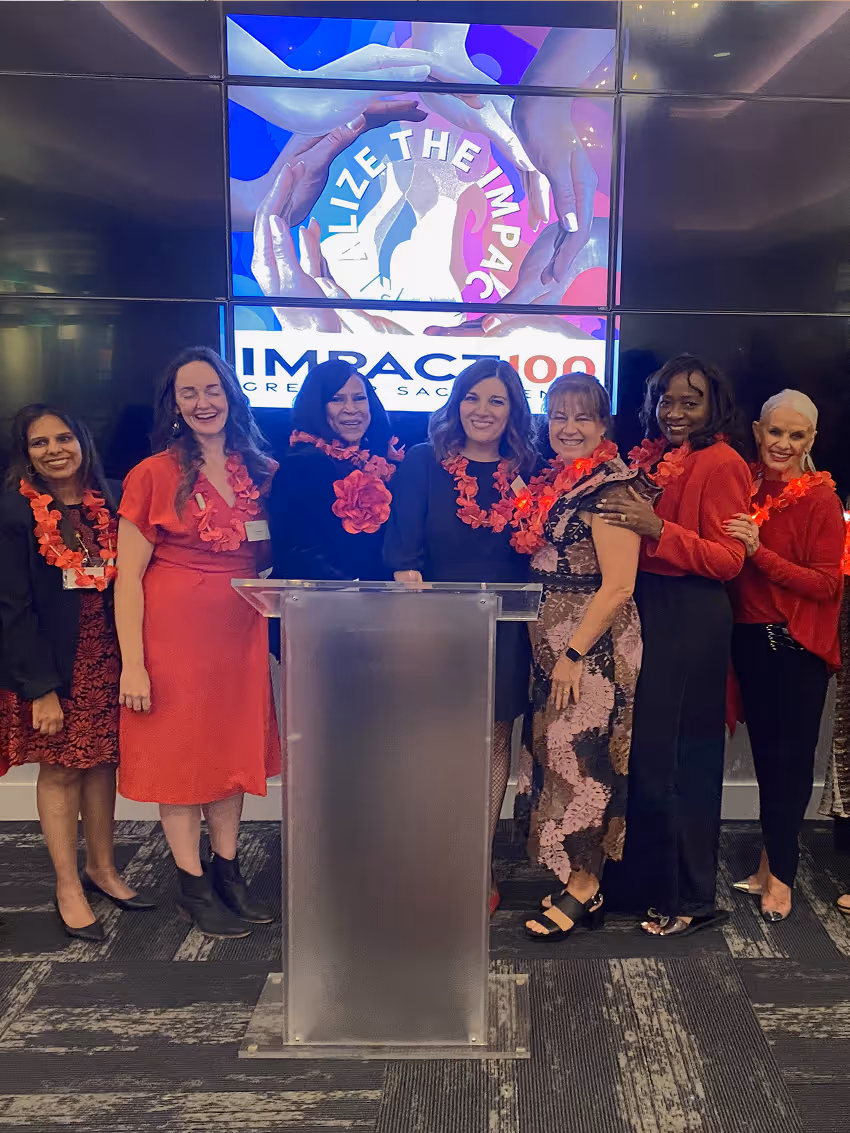 Group of women wearing red leis standing behind a podium with a screen showing Impact100 logo.