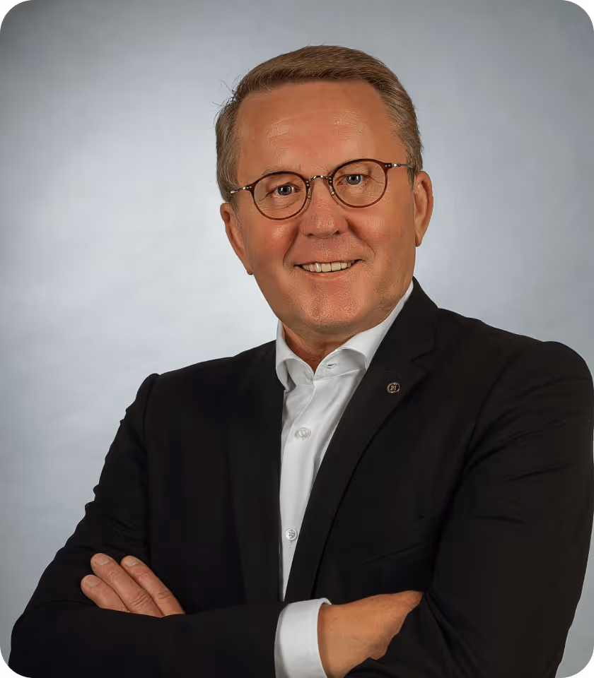 Mann in Anzug mit Brille lächelt und verschränkten Armen vor grauem Hintergrund.