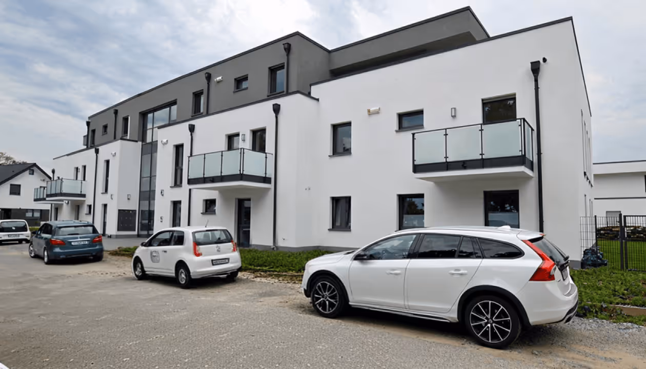 Modernes Mehrfamilienhaus mit weißen Wänden, mehreren Balkonen und geparkten Autos auf einer asphaltierten Straße.