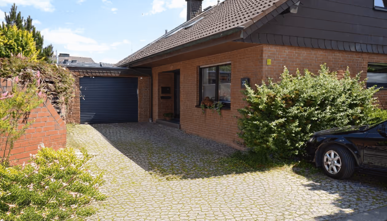 Einfamilienhaus mit rotem Klinker, gepflasterter Einfahrt, Garage und schwarzem Auto im sonnigen Garten.
