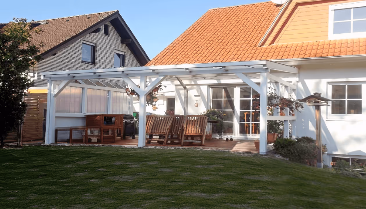 Weiße Pergola auf Terrasse eines Hauses, umgeben von grüner Rasenfläche, mit Holzmöbeln und hängenden Blumen.