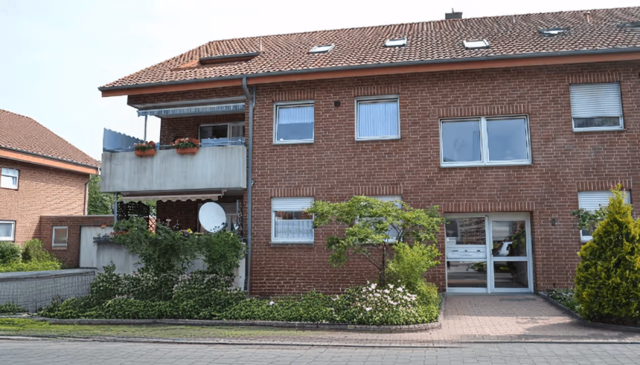 Zweistöckiges Wohnhaus aus rotem Backstein mit Balkon, Dachfenstern und gepflegtem Vorgarten.
