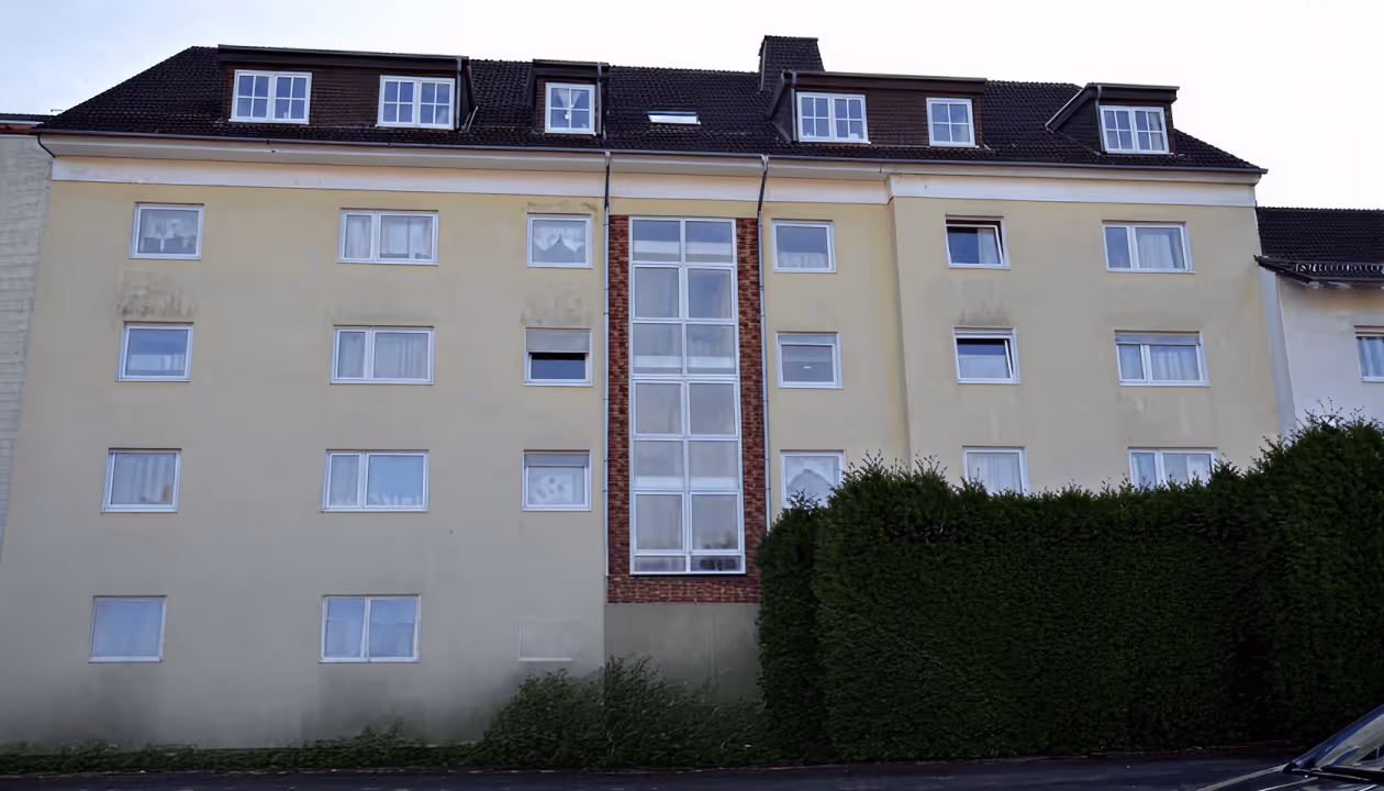 Wohnhaus mit gelber Fassade, großen Fenstern, hellem Treppenhaus und grünem Heckenpflanzen am Rand.