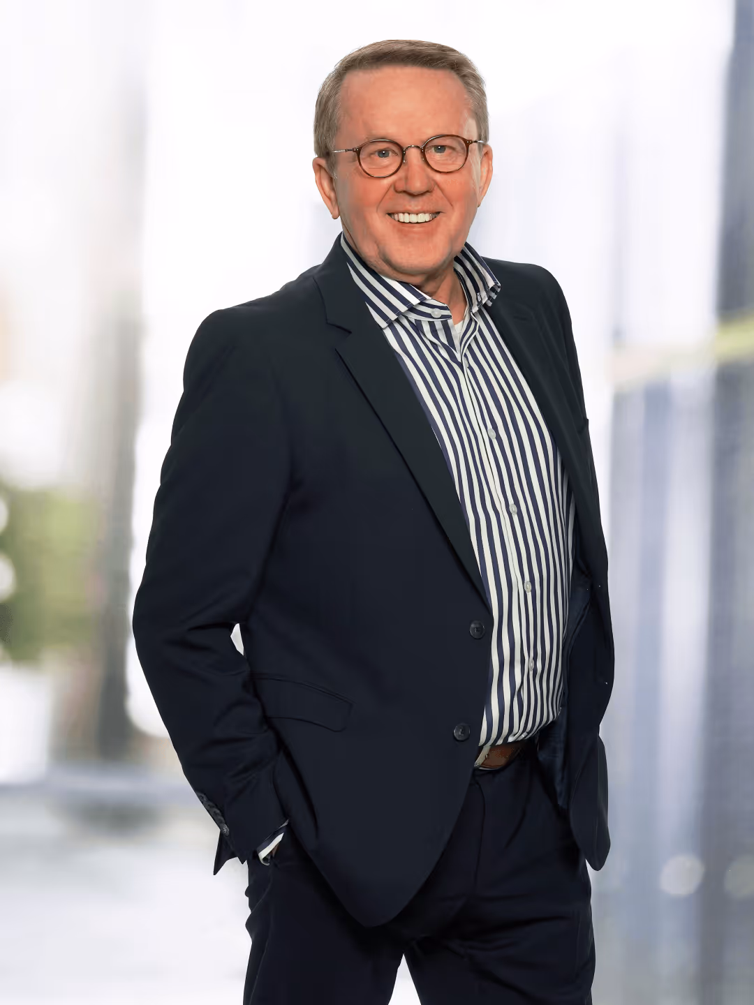Mann im Anzug mit gestreiftem Hemd, lächelnd und mit Brille, steht vor verschwommenem Hintergrund.