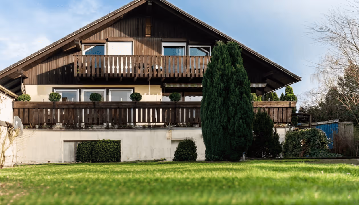 Zweistöckiges Holzhaus mit Balkon, grüner Rasen, konischer Baum und klare Himmel im Hintergrund.