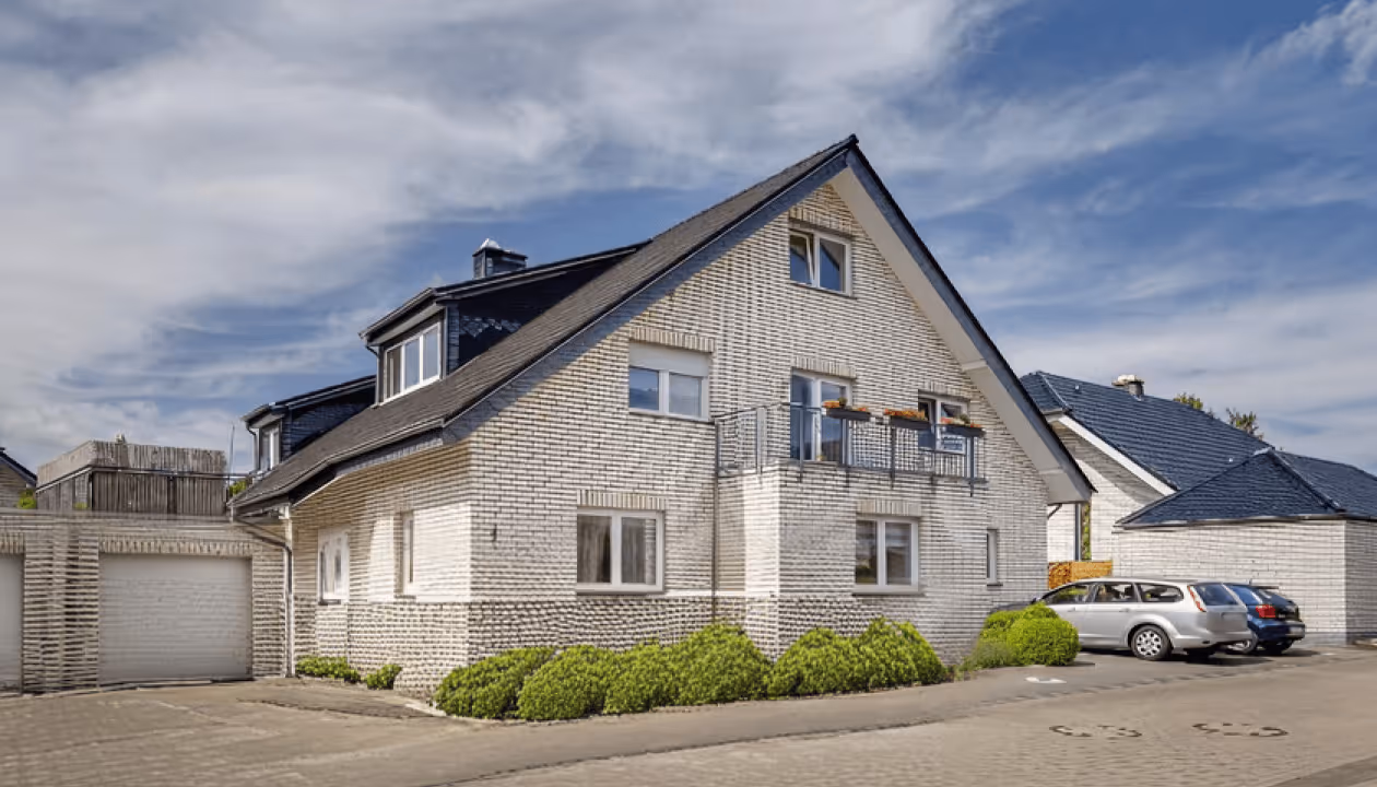 Zweistöckiges Einfamilienhaus mit weißen Klinkern, blauer Dachschräge, kleinem Balkon und geparkten Autos davor.