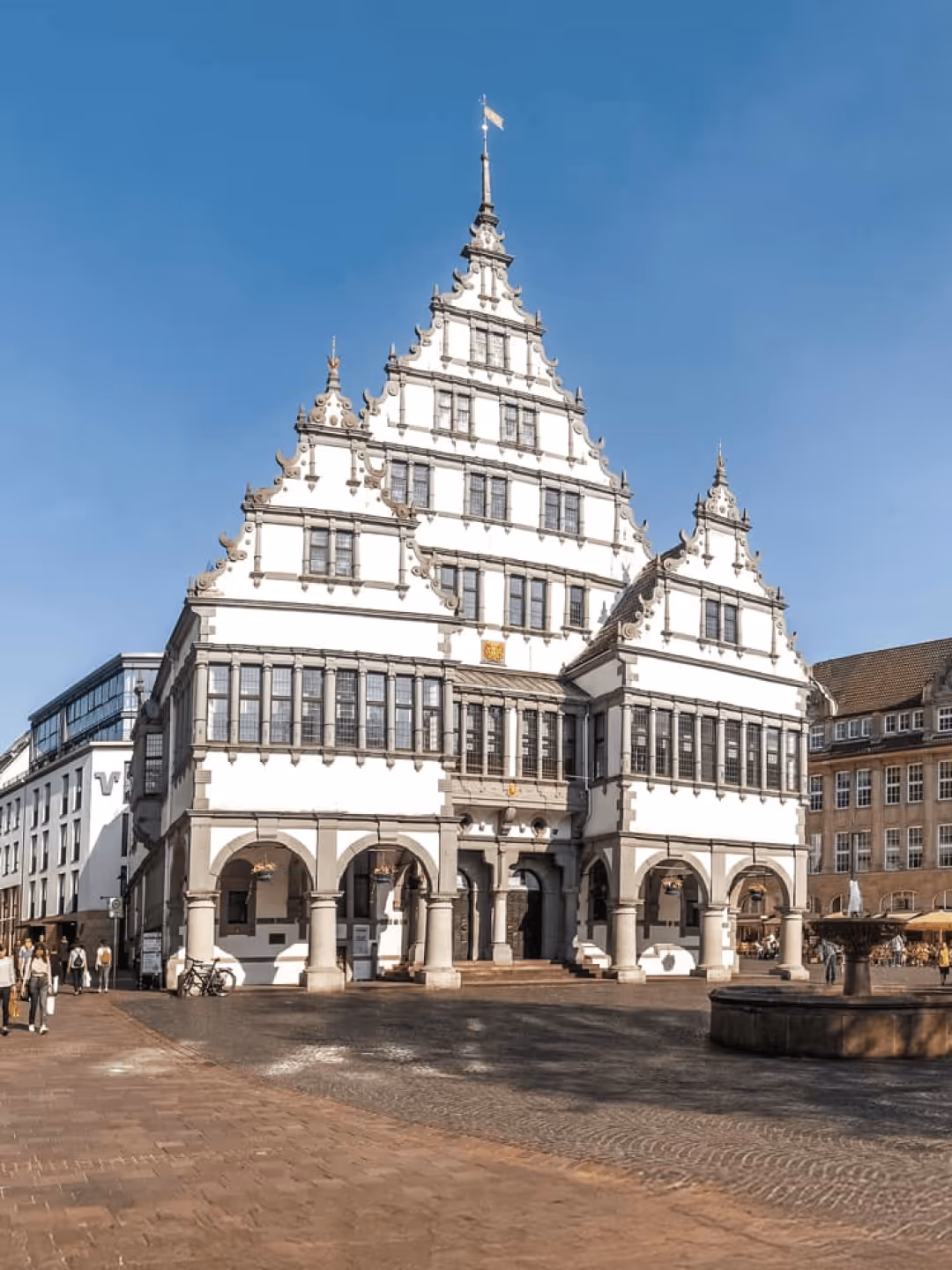 Historisches mehrstöckiges Gebäude mit Giebeln und Säulen, sonniger Tag, blauer Himmel, Menschen spazieren auf dem Platz.